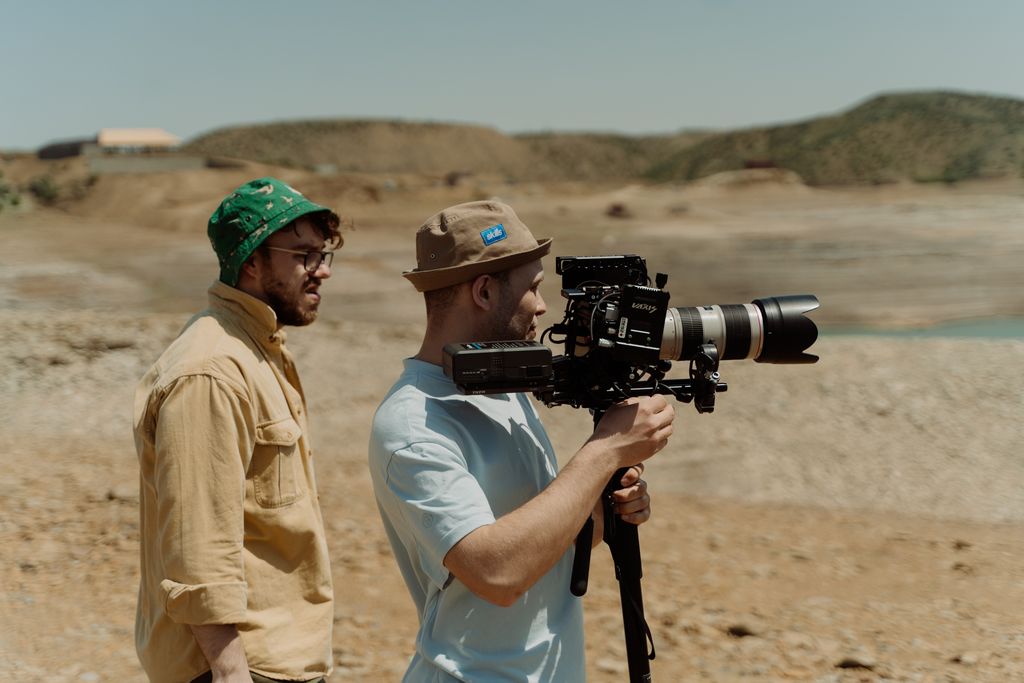 Two filmmakers operating a professional video camera outdoors in a rocky landscape.